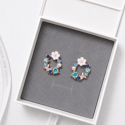 Blue floral crystal stud earrings with pearl accents and rose gold details displayed in a luxury gift box.